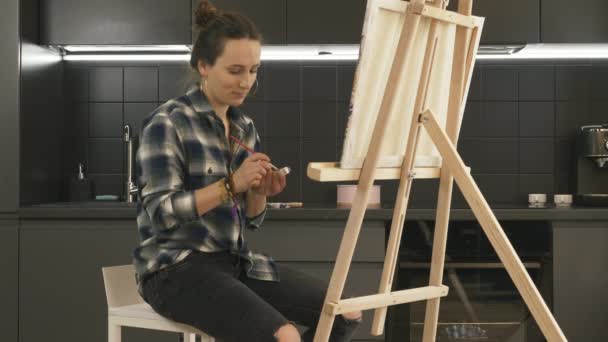 여성 미술가는 집 뒤에 부엌 이 있는 캔버스 위에 그림을 그리기도 합니다. 재능있는 젊은 여자가 이젤 근처 의자에 앉아서 아크릴 페인트를 집에서 사용하여 그림을 그리는 모습 — 비디오
