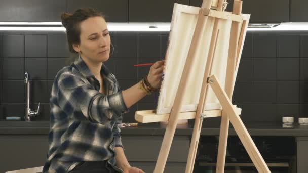 Tehetséges lány festi vászonra a modern konyhában otthon. Portré fiatal női művész festés vászonra segítségével vékony ecset. Női arc rajzol egy képet az állványra. Tehetség. Inspiráció