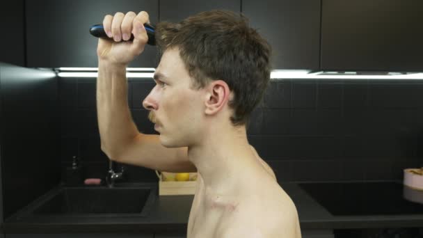Młody facet ze stylowymi wąsami golący głowę elektrycznym przycinaczem w domu w kuchni. Mężczyzna zostaje w domu podczas kwarantanny i goli sobie głowę golarką elektryczną. — Wideo stockowe