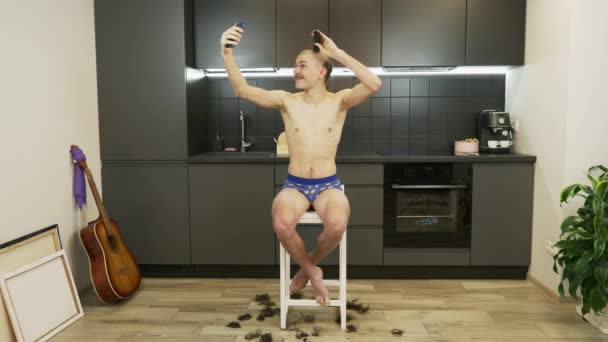 Grappige man zit in de keuken, scheert zijn hoofd door elektrisch scheerapparaat en maakt foto 's op smartphone. Man in onderbroek scheert hoofd met elektrisch scheermes en doet selfies. Zelfisolatie op quarantaine — Stockvideo