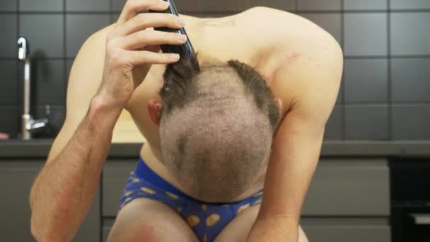 Alsógatyás fiatal fehér férfi borotválja a fejét elektromos vágóval otthon, elszigetelve a karantén alatt. Egy fickó borotvával borotválja a fejét, mielőtt bevonul a seregbe.. — Stock videók