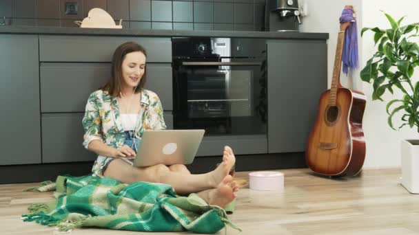 Mujer con auriculares escuchando música usando un portátil sentado en el piso de la cocina. La chica se queda en casa durante la cuarentena del coronavirus. Mujer relajándose en la cocina — Vídeos de Stock