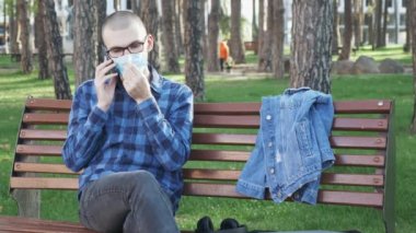 Güneşli bir günde, şehir parkında konuşan, tıbbi maske takmış beyaz bir gencin portresi. Parkta bankta oturan ve telefonda konuşan, koruyucu maskeli bir erkek.