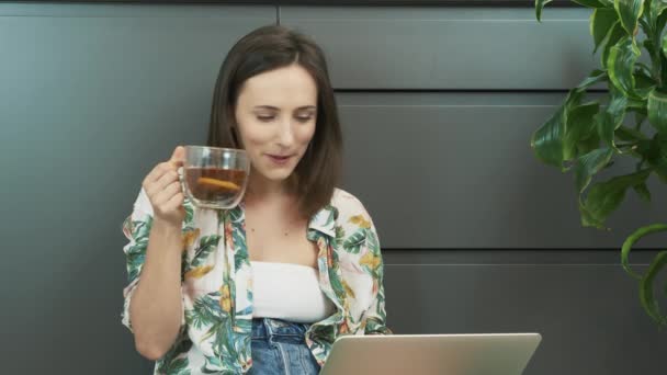 Jong lachend meisje dat thuis aan laptop werkt en thee drinkt. Een vrouw werkt op een laptop op een afgelegen werkplek. Vrouwelijke kijken op laptop scherm, zoeken naar informatie op internet, kijken naar online training — Stockvideo