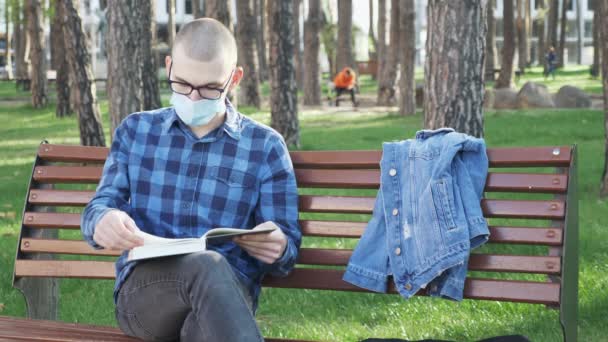 Millennial masculino em máscara facial médica ler livro no parque da cidade durante a quarentena. Homem caucasiano em óculos e máscara protetora descansando e relaxando no parque com árvores verdes — Vídeo de Stock