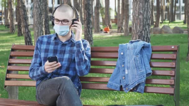 Человек в защитной медицинской маске и наушниках, слушающий музыку, сидя в городском парке на скамейке запасных. Молодой парень в очках и наушниках слушает музыку и отдыхает в парке — стоковое видео