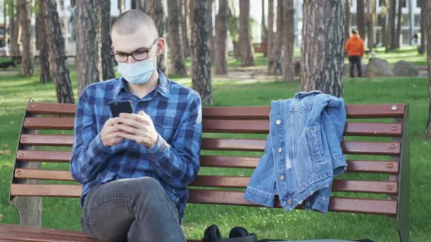 Tipo con máscara médica protectora con teléfono inteligente en las manos sentado en el banco en el parque de la ciudad. Joven hombre calvo usando el teléfono mientras descansa en la zona del parque rodeado de árboles — Vídeos de Stock