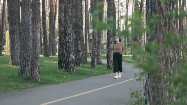 Chica morena europea en máscara médica protectora caminar solo en el parque de la ciudad. Mujer joven con mascarilla médica caminando por el parque y respirando aire fresco durante la epidemia de coronavirus covid-19 — Vídeos de Stock