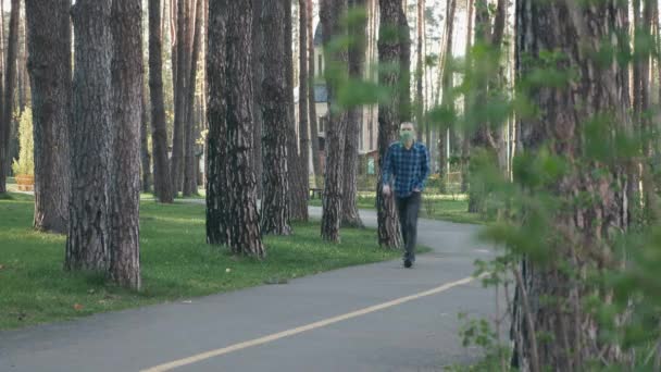 Ung man i skyddande medicinsk mask gå ensam i stadsparken. Vit man i medicinsk ansiktsmask går i parken och andas frisk luft under coronavirus covid-19 epidemi — Stockvideo