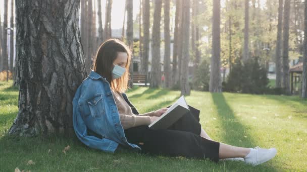 Jeune femme seule dans un masque médical protecteur livre de lecture dans le parc de la ville vide pendant la pandémie de coronavirus covid-19. Hipster fille en tenue décontractée et masque livre de lecture et de repos dans le parc sur le coucher du soleil — Video