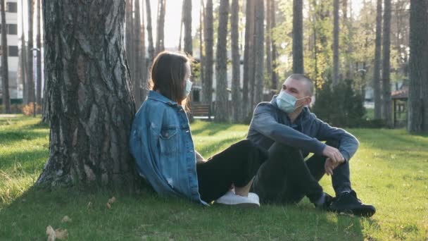 Щаслива пара в захисних медичних масках сидить на траві в парку і розмовляє, розважається, відпочиває під час пандемії ковадла-19. Молода дівчина і хлопчик відпочивають у парку на заході сонця. Карантин коронавірусу — стокове відео