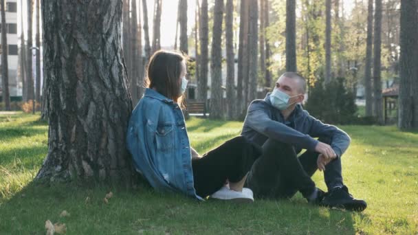 Glückliches Paar in medizinischen Schutzmasken sitzt auf Gras im Park und redet, hat Spaß und ruht sich während der Covid-19-Pandemie aus. Junge Mädchen und Jungen entspannen sich im Park bei Sonnenuntergang. Coronavirus-Quarantäne — Stockvideo