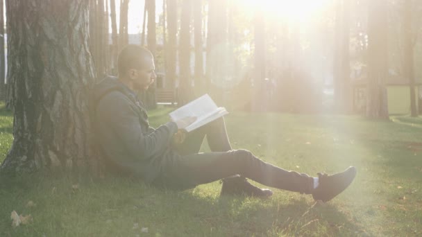 Giovane bell'uomo seduto sull'erba nel parco al tramonto e leggere libro. Un tizio che legge libri e si riposa nel parco al tramonto. Concetto di persone felici e stile di vita — Video Stock