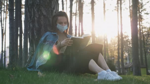 美しい夕日で公園の芝生の上に座っている間、保護医療用マスクの女の子はリラックスして本を読んでいます。検疫中に公園で本を読んで医療面マスクでかわいい女性の肖像画 — ストック動画