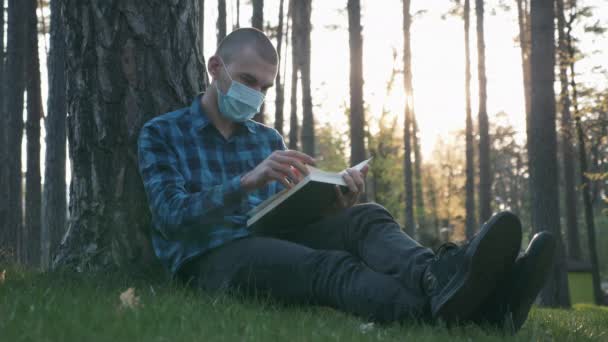 美しい夕日の公共公園で草の上に座っている間、保護医療用マスク読書本や休憩中の男。検疫中に公園で本を読んで医療面マスクの男性の肖像 — ストック動画