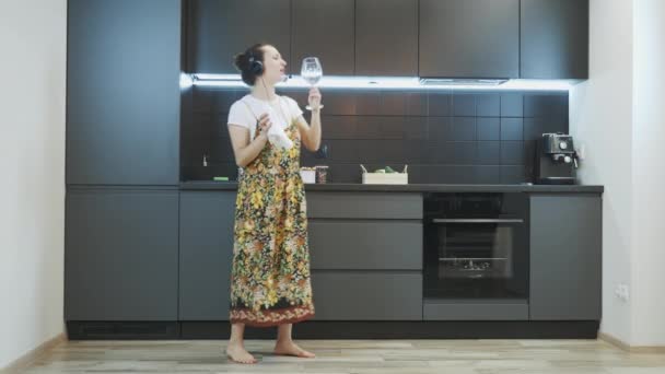 Mujer joven o ama de casa limpia el vidrio con una toalla después de lavar los platos. Feliz chica sonriente en auriculares limpiando platos después de la cena, cantando y bailando en la cocina moderna. Concepto de trabajo doméstico — Vídeo de stock