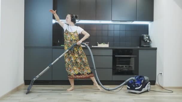 Młoda szczęśliwa kobieta sprząta dom odkurzaczem, słucha muzyki w słuchawkach, śpiewa i tańczy. Nastolatka bawiąca się w domu odkurzając podłogę w kuchni. Koncepcja prac domowych — Wideo stockowe