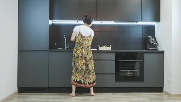 Mulher sorridente limpando cozinha moderna, dançando e ouvindo música em fones de ouvido. Mulher feliz em casa casual desgaste se divertindo e limpeza casa, cantando e dançando — Vídeo de Stock