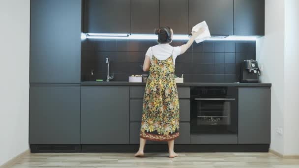 La mujer limpia muebles en la cocina. Joven mujer caucásica o ama de casa limpiando la cocina, escuchando música en auriculares y bailando. Chica usando trapo blanco para limpiar muebles de cocina — Vídeo de stock