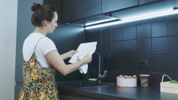 Egy fiatal nő letörli a tányért fehér törölközővel a modern konyhában. Házimunka és takarítás. A lány törülközővel törli az edényeket. A nő takarít a konyhában. — Stock videók
