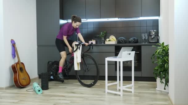 Fille faisant des exercices sportifs sur le vélo entraîneur et l'eau potable ou isotonique de la bouteille pendant l'entraînement. Femme vélo intérieur sur vélo stationnaire pendant l'auto isolement et la distance sociale — Video