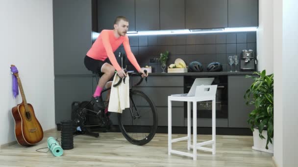 Allenamento sportivo uomo su ciclismo allenatore stazionario a casa durante la pandemia coronavirus per distanza sociale auto isolato in quarantena. Ragazzo in jersey rosa pedalando su bicicletta fissa indoor — Video Stock