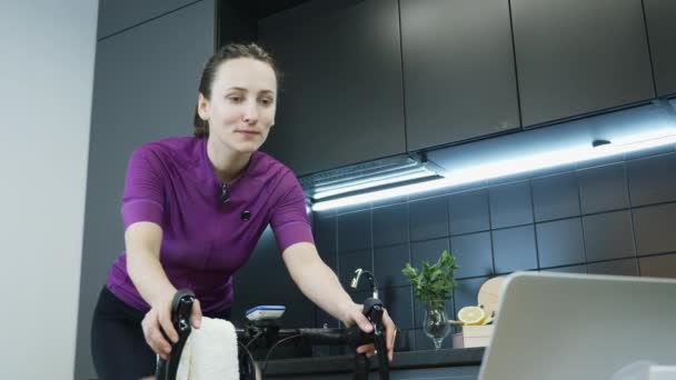 コロナウイルスのパンデミックの間に家にいる屋内スマート自転車トレーナーに乗るサイクリングアパレルの若い女性の肖像画。若いです笑顔女の子ペダリングオン自転車やオンラインサイクリングトレーニングを見て — ストック動画
