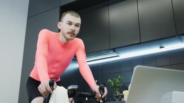 Retrato de un joven en jersey de ciclismo rosa entrenando en un entrenador de bicicletas estacionario inteligente y viendo carreras de ciclismo en línea en un ordenador portátil. Chico paseo en bicicleta estacionaria quedarse en casa en cuarentena — Vídeos de Stock