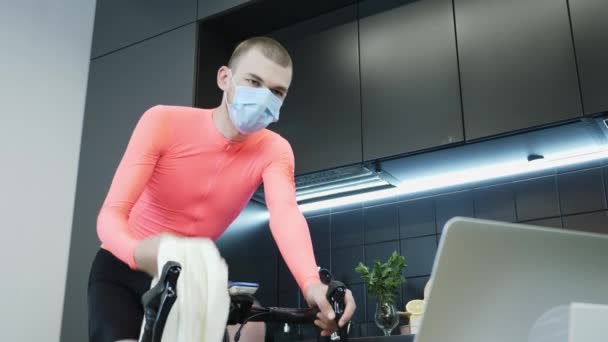 Профессиональный велогонщик в защитной медицинской маске для лица педалирует и тренируется на умных стационарных тренажерах по велоспорту, наблюдая за онлайн-тренировками на ноутбуке. Концепция самоизоляции — стоковое видео