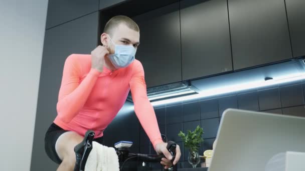 Парень снимает защитную медицинскую маску для лица и улыбается после тренировки по физкультуре на умном стационарном велосипедном тренере, оставаясь в самоизоляции на коронавирусном карантине. Концепция Stay Home — стоковое видео