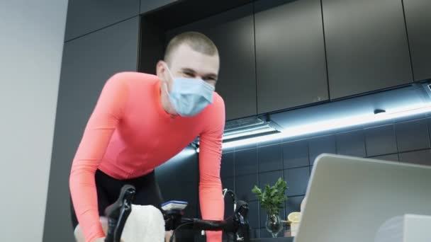 Jovem homem esportivo em roupas de ciclismo rosa pedalando intensamente em bicicleta estacionária inteligente e participando de corridas de bicicleta on-line ficar em casa para auto-isolamento durante coronavírus covid-19 pandemia — Vídeo de Stock