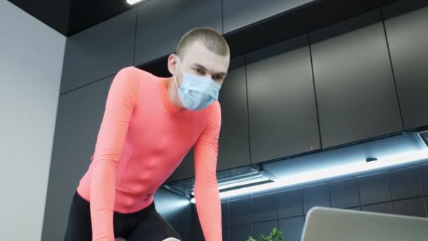 Retrato del atleta joven en forma en el entrenamiento de máscara médica protectora en casa en bicicleta estacionaria durante la cuarentena. Tipo deportivo haciendo ejercicios de cardio en entrenador de ciclismo inteligente en casa en la cocina — Vídeo de stock