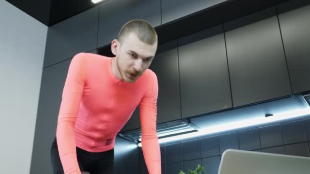 Young man in sportswear cycling and pedaling indoor on smart stationary bicycle trainer, doing cardio exercises and watching online fitness trainings on laptop computer staying home during quarantine — Stock Video