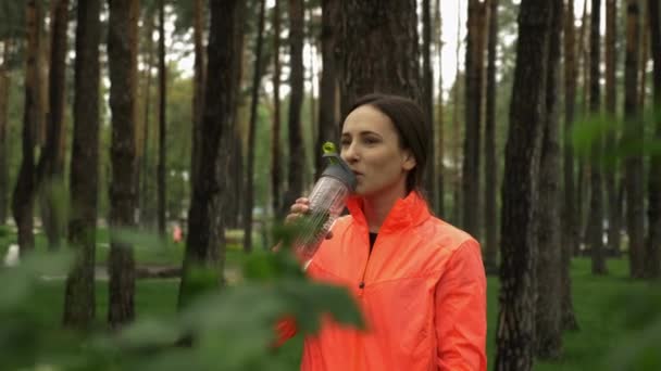 Portrait d'une belle athlète boit de l'eau de bouteille dans le parc entouré d'arbres verts. Brunette fitness femme boit de l'eau après l'exercice de course sur l'air frais dans le parc de la ville — Video