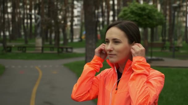 Happy sport woman put on earphones and use smartphone before running training standing in empty city park on rainy day. Portrait of fitness female in park wearing earphones before jogging exercises — Stock Video