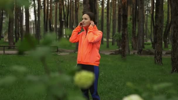 Mulher fitness coloca fones de ouvido e preparar seu smartphone para o treinamento de corrida. Esporte atleta feminina usando fones de ouvido e usando o aplicativo de treinamento no smartphone antes de correr no parque da cidade chuvosa de manhã — Vídeo de Stock