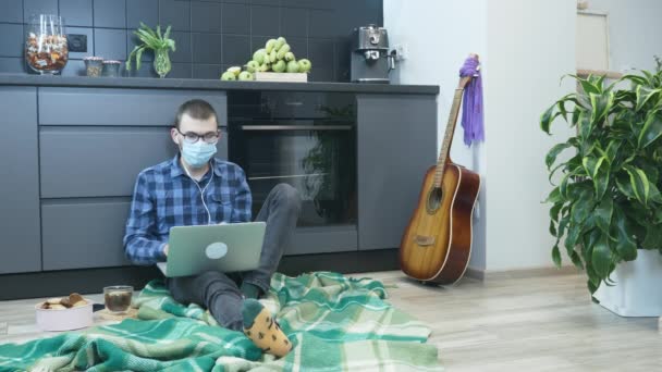 Człowiek w słuchawkach i masce medycznej słuchający muzyki za pomocą laptopa siedzącego na podłodze w kuchni. Mężczyzna przebywa w domu podczas kwarantanny koronawirusowej — Wideo stockowe