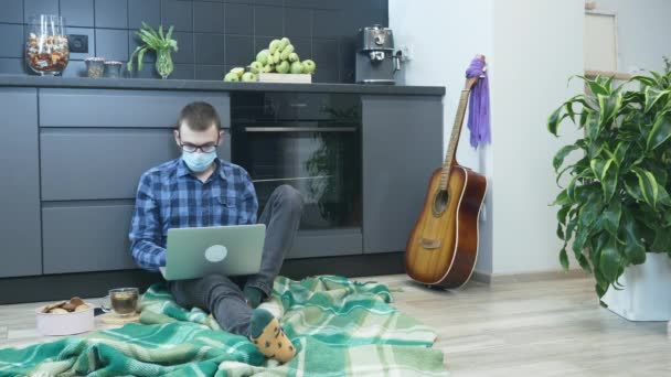 Człowiek w masce medycznej pracujący na laptopie w odległym miejscu pracy. Mężczyzna korzystający z laptopa w biurze podczas samoizolacji w domu. Mężczyzna siedzi na podłodze w kuchni i pracuje na laptopie — Wideo stockowe