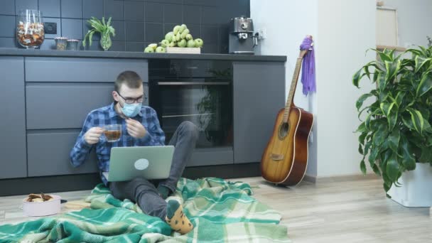 戴防护医疗面罩的学生坐在地板上用手提电脑喝茶。男人在工作时摘下蓝色的医疗面罩，在笔记本上喝茶 — 图库视频影像