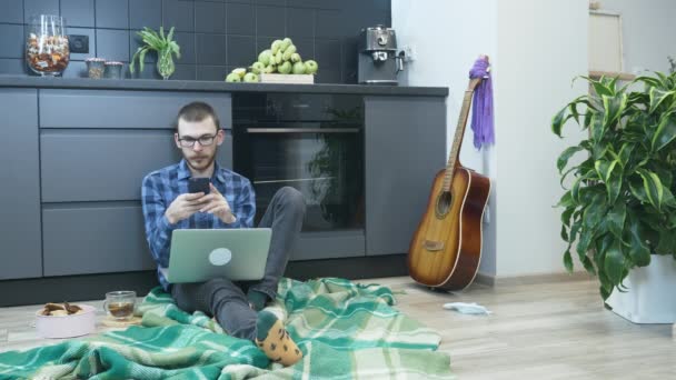 年轻的嬉皮士戴眼镜，穿着休闲装和可笑的袜子坐在厨房的地板上，用智能手机，在社交网络上与朋友聊天，回复邮件，看新闻 — 图库视频影像