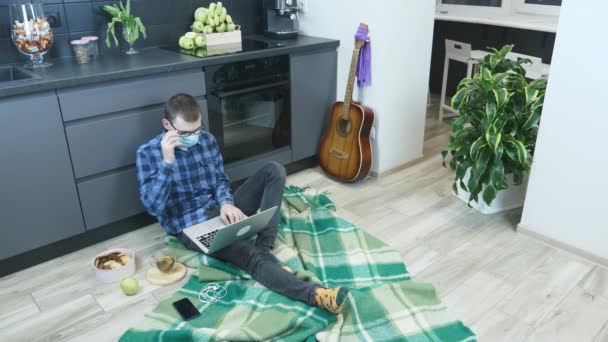 Estudante sentado no chão na cozinha e usando computador portátil para projeto remoto. Hipster homem tira óculos e máscara médica protetora após o trabalho duro no caderno. Trabalhando em casa — Vídeo de Stock