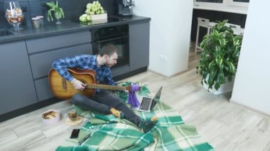 Erkek gitarist çevrimiçi gitar dersleri izliyor ve yeni akorlar çalmaya çalışıyor. Adam mutfakta yerde oturuyor ve online öğretmenle akustik gitar çalmayı öğreniyor. Online gitar dersleri