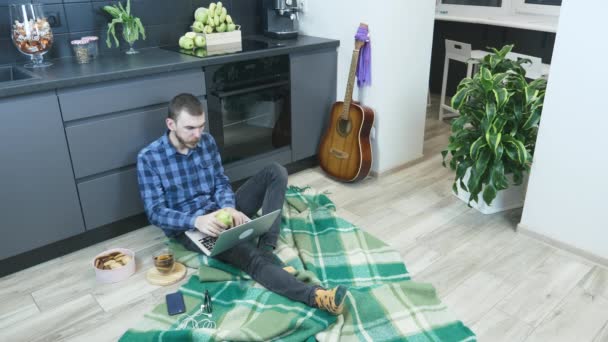 年轻的留胡子男子在家里做远程项目时使用笔记本电脑。带着电脑的男人，坐在厨房的地板上，一边吃苹果一边玩。在家中检疫时工作 — 图库视频影像