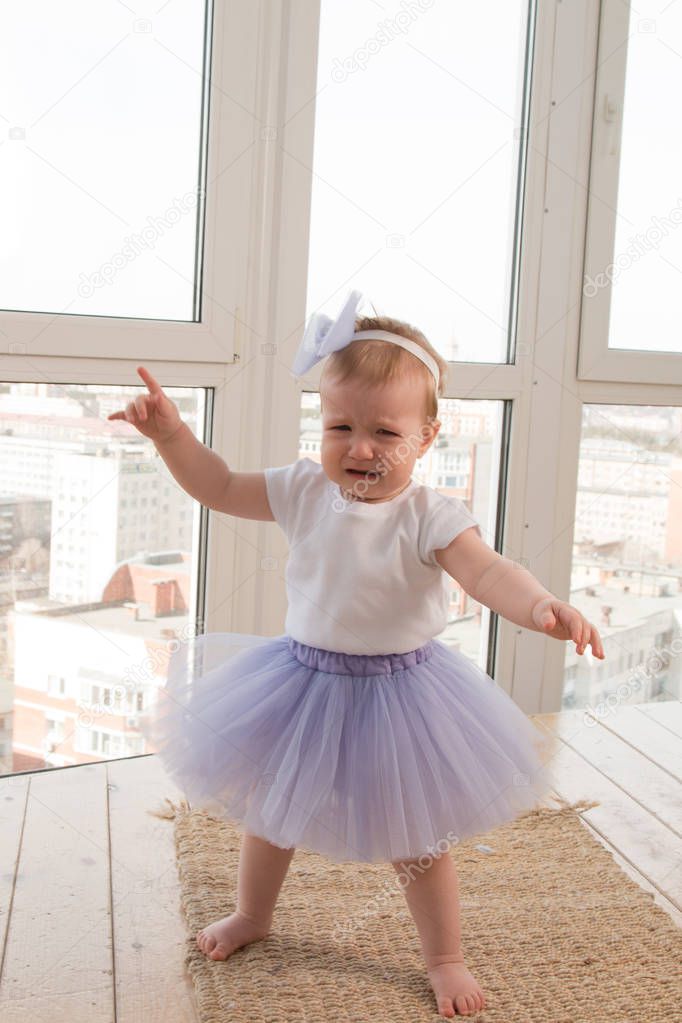 A cute little girl in a lilac ballet tutu stands by the window and cries