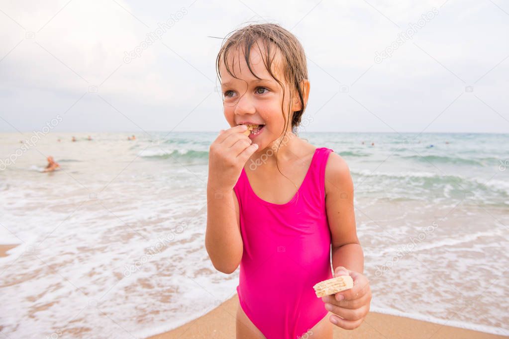 The girl in the pink bathing suit standing on the beach and eating waffle