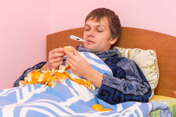 Больной ребенок с термометром во рту лежит в постели и ест мандарины — стоковое фото
