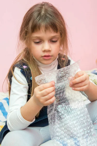 Chica de cinco años sentada en la cama y come burbujas paquete de embalaje — Foto de Stock