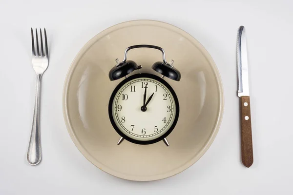 On the plate is an alarm clock, lying next to a knife and fork, white background, top view — Stock Photo, Image