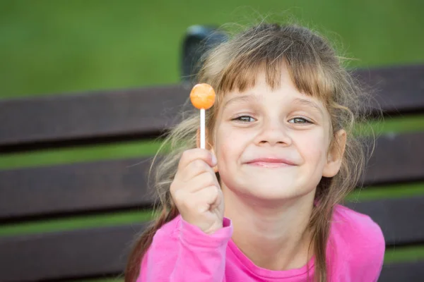 Portrait of a six-year-old cheerful girl who holds a lollipop in her hand and looks pleased at the frame — Stock Photo, Image