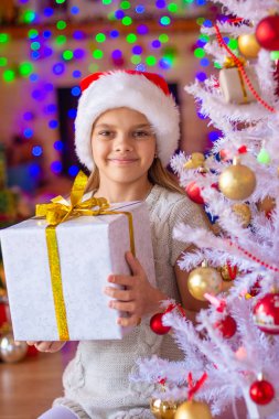 Kız elinde hediyelerle Noel ağacının yanında oturuyor.